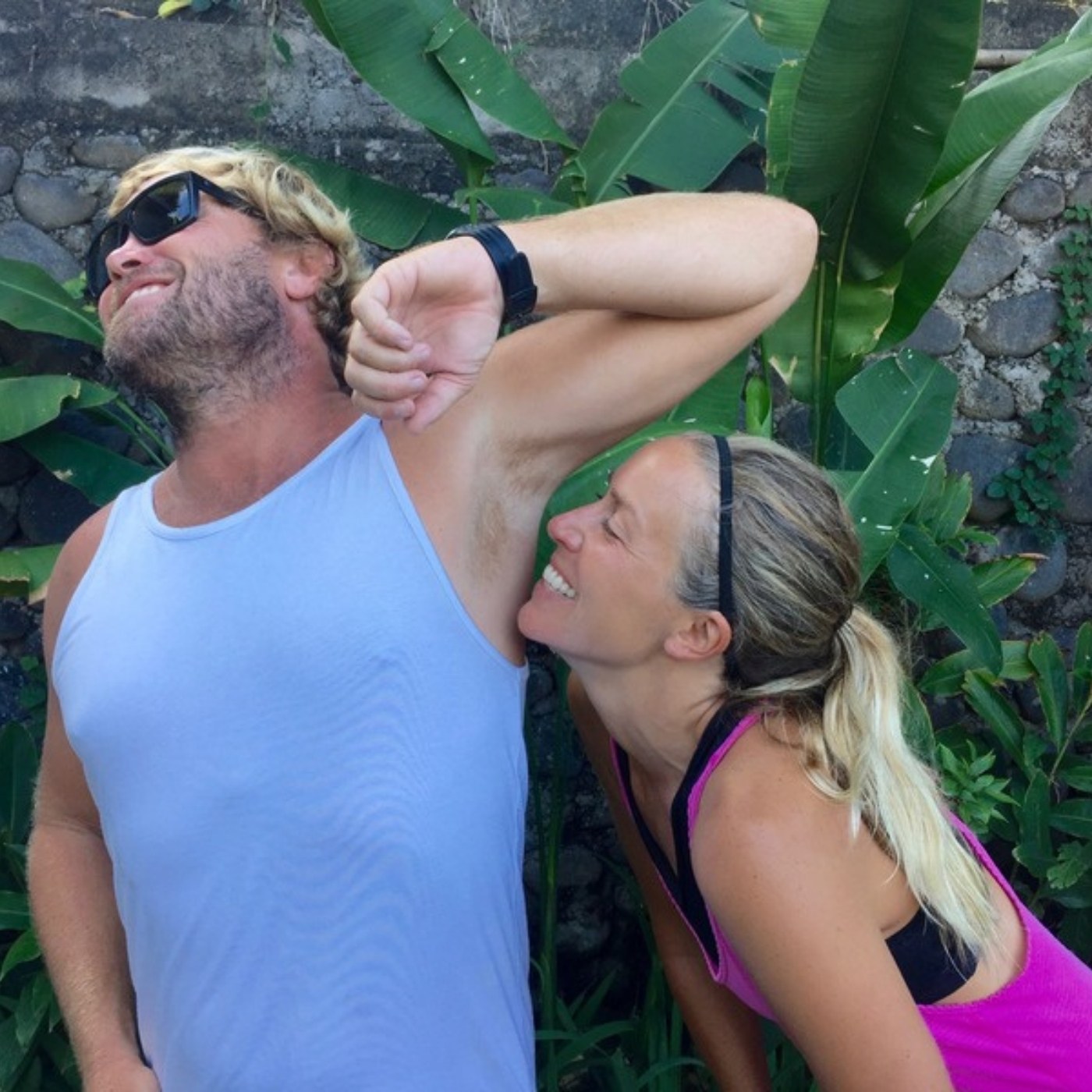 A man in a white singlet lifting his arm while a laughing woman leans in to smell his underarm, tropical leaves behind them