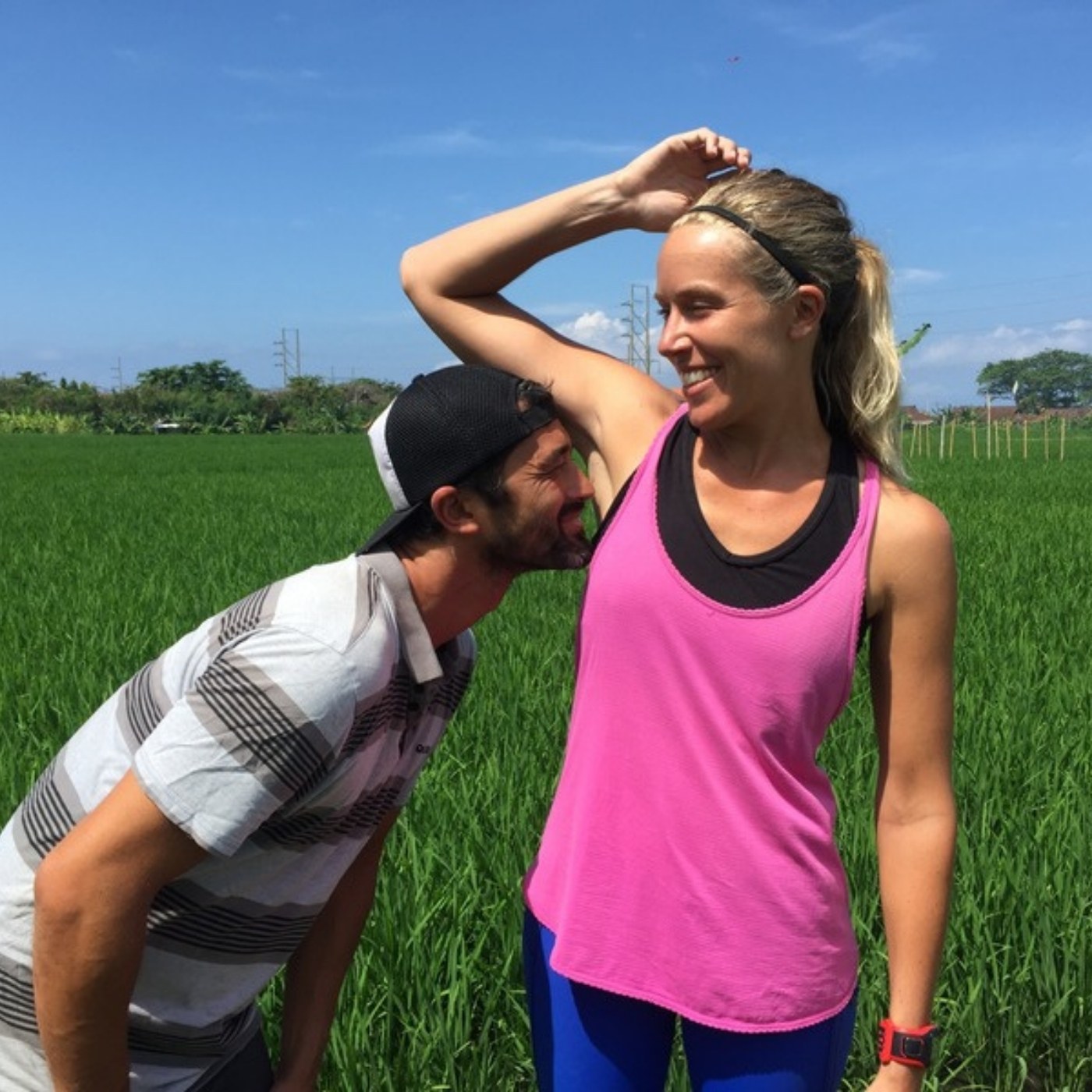 A couple in activewear standing in a green paddy field, one lifting an arm while the other leans in to smell it