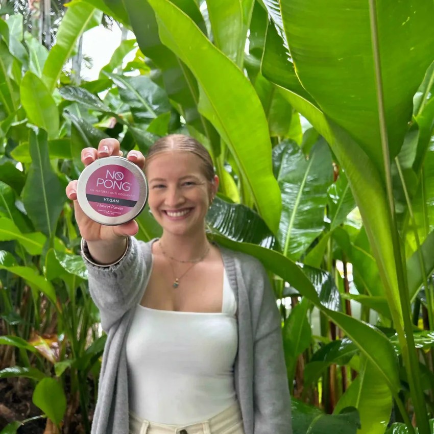 Woman smiling among tropical leaves, holding up a No Pong tin